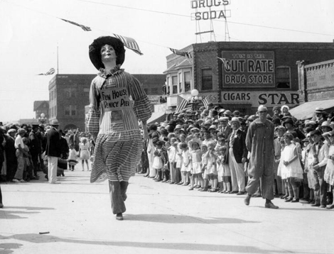 Mardi Gras parade of 1935 or 6 at Venice Beach showing a crowd on both sides of the street as a marcher wearing a costume from "Fun House Venice Pier" marches by.
