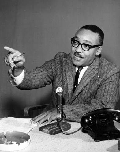 Black and white photo of Louis Lomax sitting at a desk and speaking into a microphone.