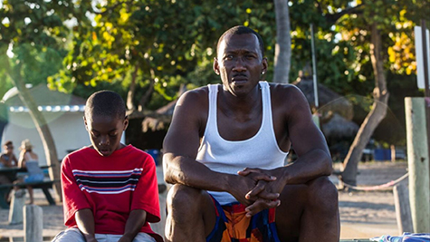 a black man sitting down, looking straight ahead and a young black boy sitting to the man's left, looking down on his lap