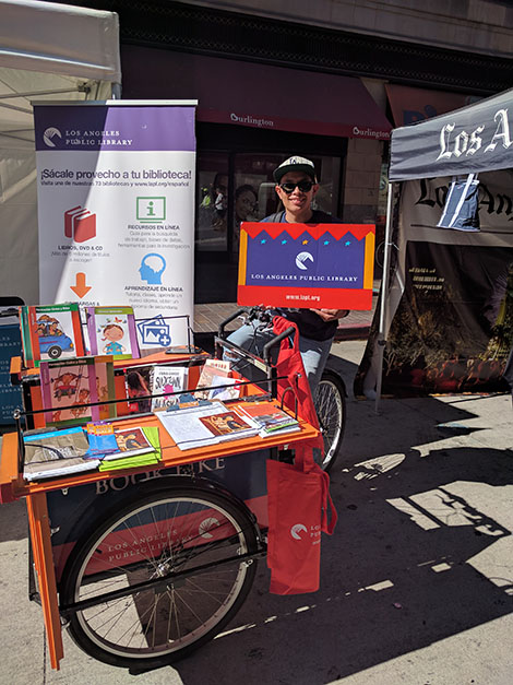 a man smiling wearing sunglasses and a baseball cap, standing in front of the library booth, holding a big library card sign.