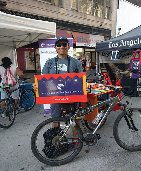 man wearing baseball cap and sunglasses holding a library card sign in front of the library booth