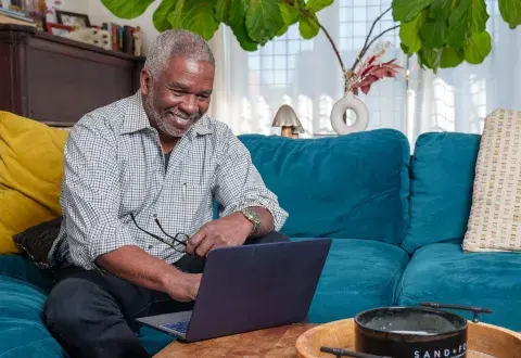 A man sits on a blue couch in a living room setting and smiles while looking at a laptop.