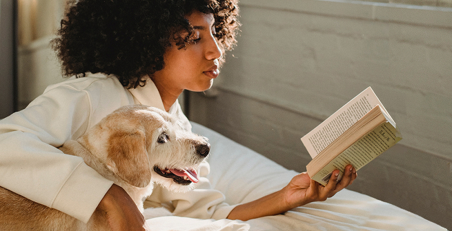 A young woman reads a book while her dog lie beside her.
