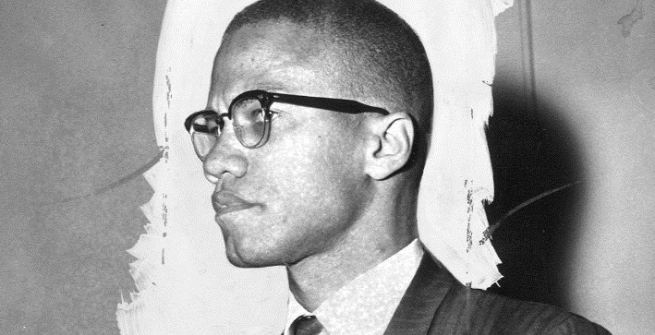 Black and white photo of Malcolm X from the side, standing at a meeting.