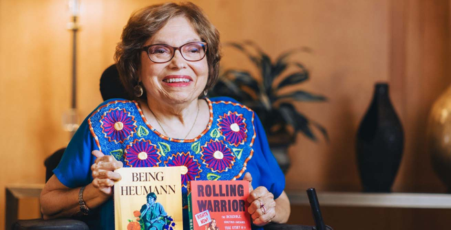 Activist Judy Heumann holding her two books, Being Heumann: An Unrepentant Memoir of a Disability Rights Activist and Rolling Warrior