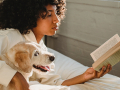 A young woman reads a book while her dog lie beside her.