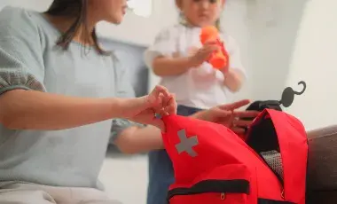 A woman packs a red backpack with a medical cross on it while a child holds a water bottle
