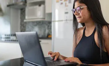 A woman with glasses and medium-length hair is seated at a desk at home, focused on using a laptop. The room has a warm and inviting atmosphere.