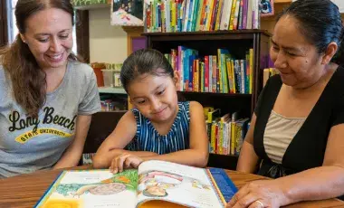 Two adults reading a book to a child. 