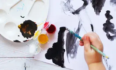 Hand holding brush painting on paper with paint palette nearby
