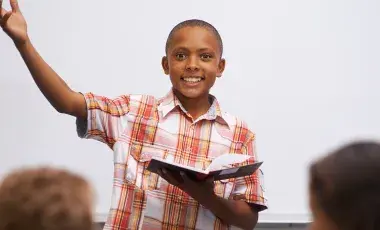 A smiling boy raises his arm as he reads a book to a classroom.
