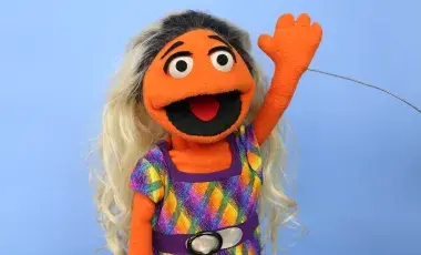 Orange Puppet girl with blond and black hair waving.