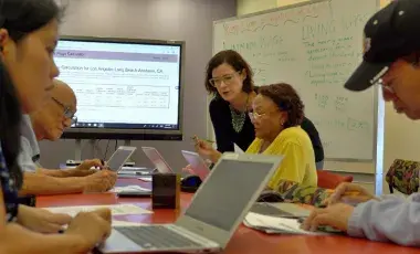 A group of adults of various ages are working collaboratively on computers in a classroom setting. 