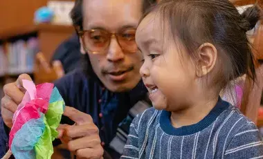 A father shows his toddler a brightly-colored toy in the library.