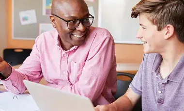 Smiling man and teenage boy talking at a desk with a laptop.