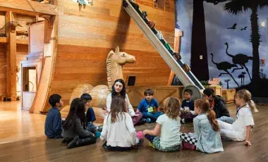 Several children and one adult interact in what looks like a stage set with a giant ark