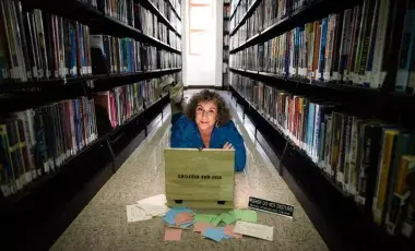 Image of artist Andy Crocker working on her computer among library bookshelves.