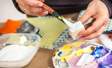 Hands painting a square accompanied by paints and craft materials.