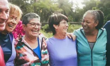 Group of older adults have arms around each other and are smiling outdoors surrounded by trees