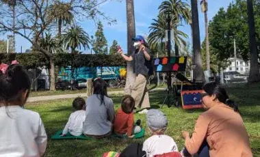 adult caregivers and toddlers seated on a grassy lawn watch a librarian doing storytime in front of trees in Echo Park