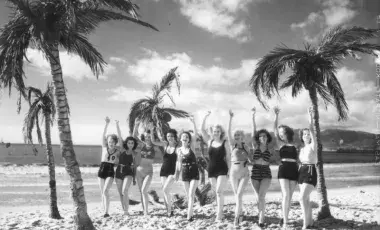 Ten young woman on a beach, surrounded by palm trees, wave at the camera. 