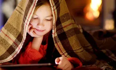 A girl reads a tablet while under a blanket