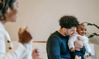 A man holds a smiling baby as a smiling woman looks on. 