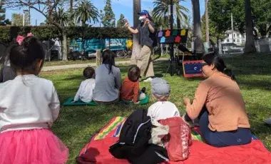 toddlers and adult caregivers seated on a grassy lawn watch a librarian leading a storytime in a park