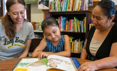 Parent and child are looking down at an open book with an Adult Literacy Coordinator helping with their reading. 