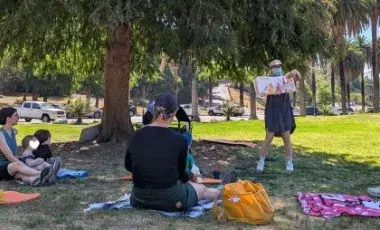 librarian reads a picturebook to a group of seated toddlers and adult caregivers in a park with trees