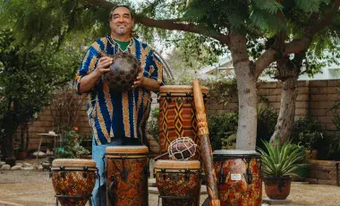 A smiling man in traditional shirt stands in front of various kinds of drums.