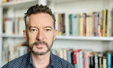 Martin Jago is pictured wearing a dark blue collared shirt, standing in front of a bookcase.