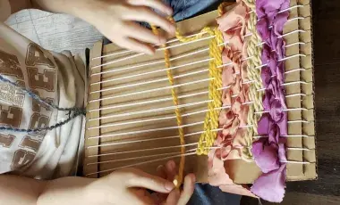 hands weaving yarns and ribbons on a cardboard loom