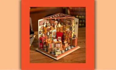 Miniature model of a room with a green and white checkboard floor, one chair, a stepladder, and many bookcases with filled with books. Tiny pictures line the walls and a miniature light fixture hangs overhead illuminating the details of the model.