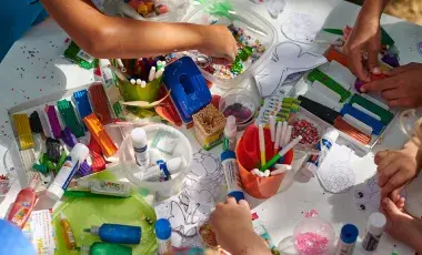 A table covered in colorful and glittery arts and crafts supplies, with various young people's hands shown reaching into the containers.