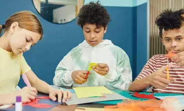 Three kids sitting at a table doing arts and crafts with paper and scissors.