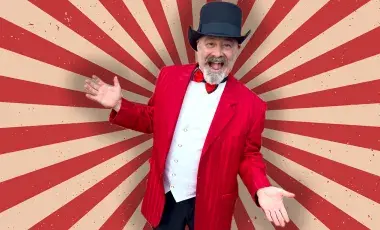 Magician wearing a black top hat and red jacket has his hands stretched out while smiling. 