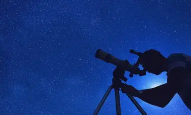 a backlit man looks through a telescope at a night sky