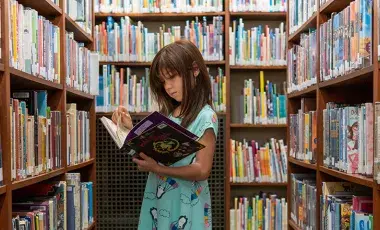 an elementary school age girl stands in a library aisle, surrounded by shelves of books, and reading an open book in her hands
