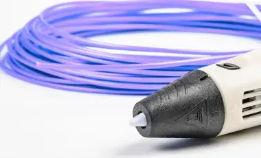 A partial view of a white 3D pen with a black tip angled at the bottom right corner and a spool of purple plasic filament behind it on a white surface.