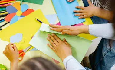 An assortment of craft supplies, paints, markers and hands with scissors cutting paper.