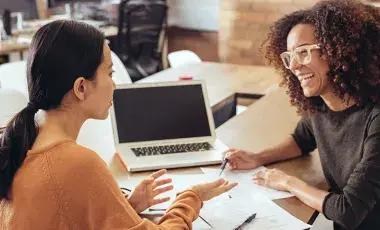 Two women sitting at a table with a laptop and papers.