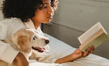 A woman reads a book on a bed with her arm around a dog.