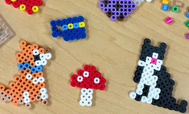 Various fuse bead creations such as a mushroom and cat are displayed with fuse bead crafting tools and unused beads.