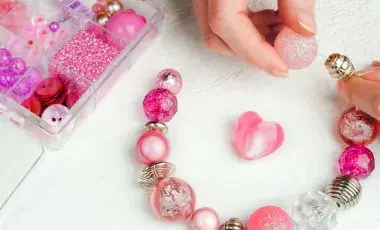 Hands assembling a pink beaded bracelet beside a tray of beads and jewelry supplies.