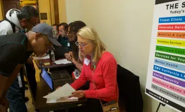 Image of a seated woman in a red sweater at a table helping a standing man with paperwork.