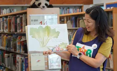 STAR Reader wearing a purple vest holds a full page spread of a picture book in surrounded by the children's library stacks. 
