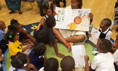 Woman holding an oversized picture book reading to a group of children.