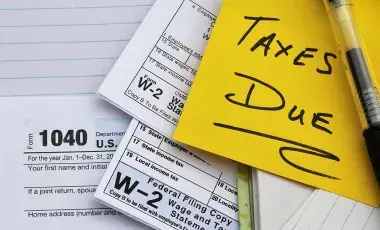 a yellow post it note with Taxes Due and three tax forms on top of a notebook