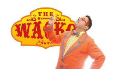 Image of a dark haired man with orange glasses and an orange jacket in front of a sign that says The Wacko Show.